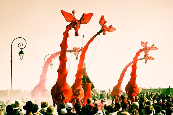 Street performers wear extraordinary red costumes, with long girafe-like necks, in a crowd of people.
