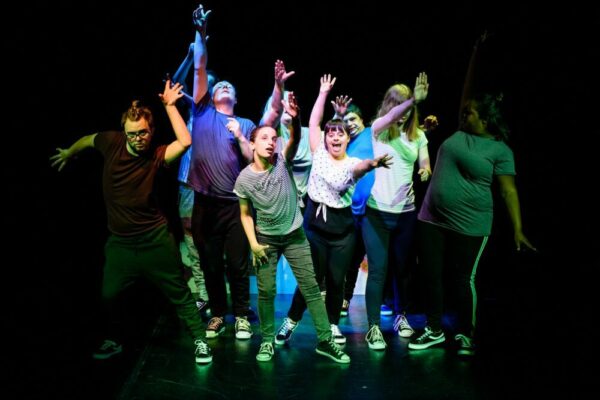 Performers pose together on a stage, reaching their hands up