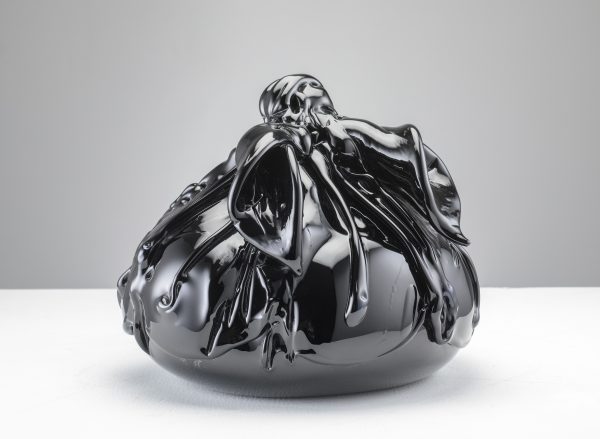 A photograph of an artwork. A round object is wrapped in shiny black material, on a white surface.