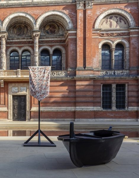 A photograph of two artworks, taken in a courtyard of the V&A gallery in London. One artwork is in the form of a fabric flag or pennant. The other is a small boat.