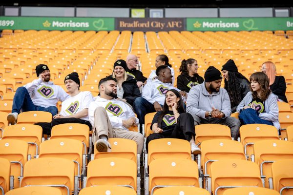 A group of people wearing Bradford 2025 branded clothing sit on seats in the Bradford City football stadium.