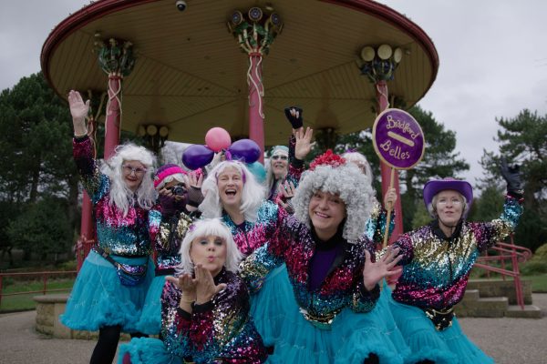 A group of people wearing colourful glittery clothes and wigs pose for the camera, smiling.
