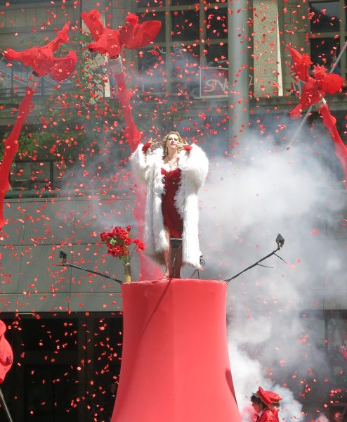 A street performer wearing a red dress and large white coat stands on top of a red platform, surrounded by red confetti.
