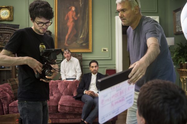 A photograph of a film set. A person with a camera faces a person with a film clapper board. Two people sit behind them ready to be filmed.
