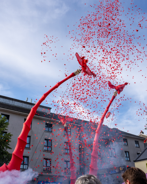 Two huge red giraffe puppets look to the sky as they are showered in red confetti
