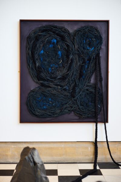 An image of an artwork on a white wall. The artwork is made up of dark rope material which flows onto the gallery floor.