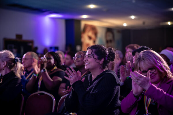 An audience are all clapping at an event. The person in the middle of the photo is a young woman smiling and clapping.