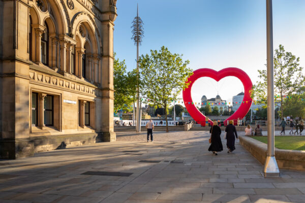 A large heart shaped red inflatable structure is in the middle of a sunny public square and two women in black outfits walk towards it.