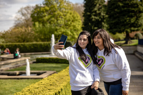Two female volunteers stand in a park in white hoodies and take a selfie of both of them