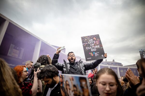 A person stands out above a crowd, with an excited smile on his face. He holds a sign that reads UK City of Culture Bid.