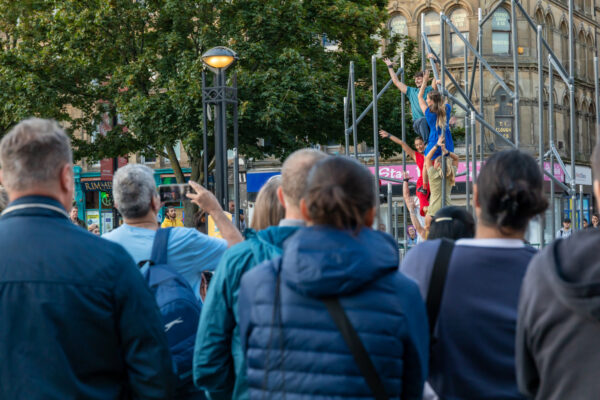 A group of men are stood together watching a performance featuring dancers climbing onto scaffolding in a busy city centre at dusk