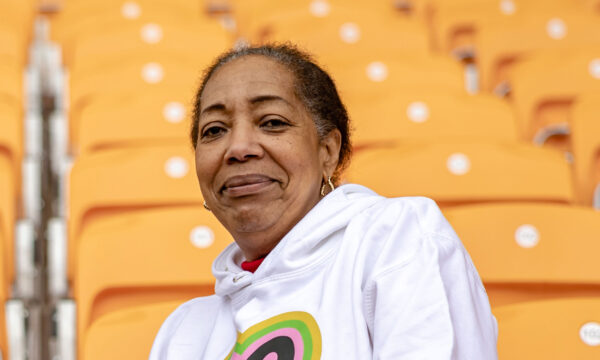 A black woman with her hair tied back sits among rows of orange chairs in a white hoodie with the Bradford 2025 logo on the front. She is smiling.