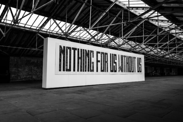 A black and white photograph of a printed artwork. In large type in all capital letters, the artwork has the words 'NOTHING FOR US WITHOUT US'