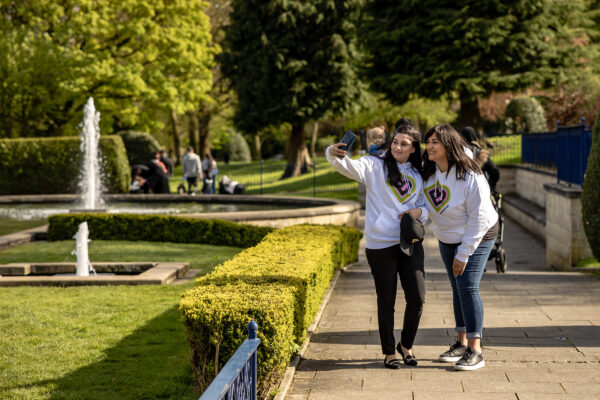 Two Bradford 2025 volunteers wear white hoodies with the BD 2025 logo. They stand in a park surrounded by greenery and take a selfie on a mobile phone.