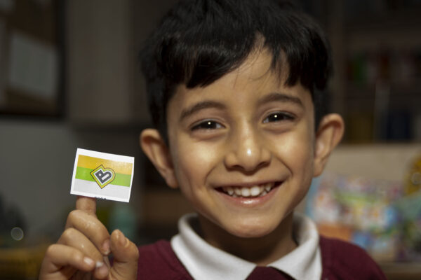 A boy smiles at the camera holding a small sticker with the Bradford 2025 logo on it.