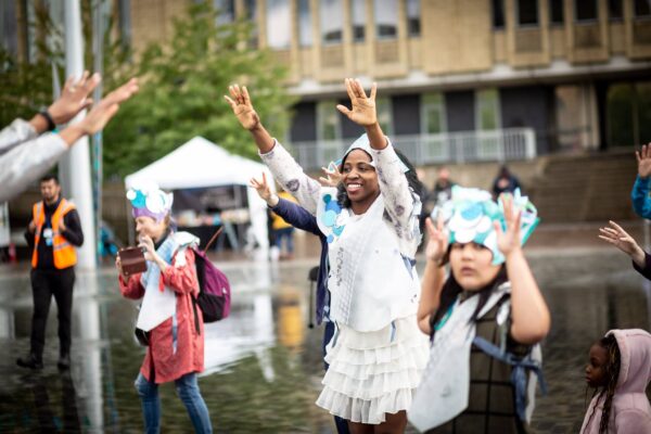 A group of adults and children wear home-made costumes in Bradford City Centre. They are reaching their hands into the air with happy expressions on their faces.