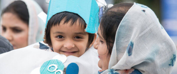 A young child wears a cardboard costume and smiles at the camera. They are being held by a woman wearing a headscarf.