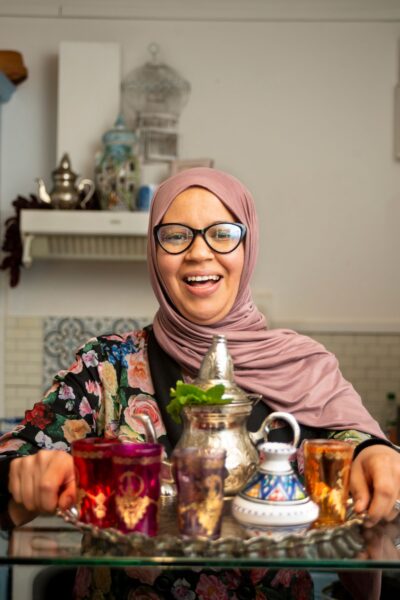 A person wearing glasses and a hijab is smiling, presenting a tray with traditional tea items. The tray holds a decorative teapot and glasses. In the background, shelves are filled with various utensils and containers, contributing to a homey atmosphere.