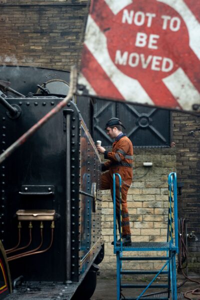 A person in overalls and a cap steps onto a vintage steam locomotive. In the foreground, a circular sign reading 