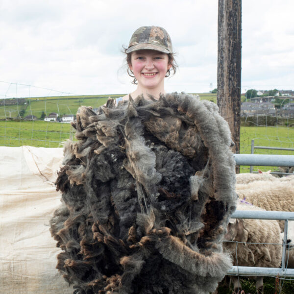 A woman holds a large sheep fleece, against a rural backdrop.