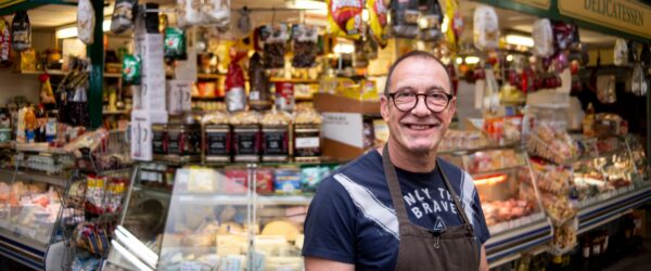 A man wearing glasses and an apron stands smiling in front of a well-stocked market stall featuring an array of continental produce.