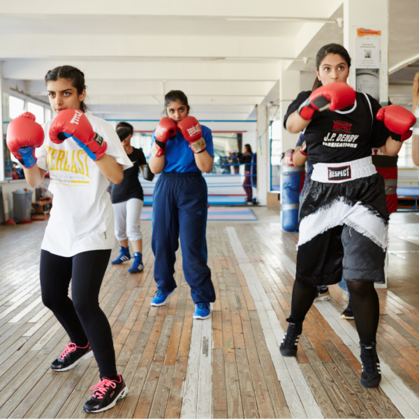 Three young people in a gymnasium practice boxing. They are wearing athletic wear and red boxing gloves.