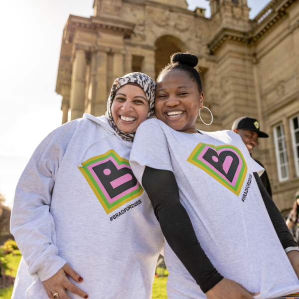 Two volunteers wearing Bradford 2025 merchandise are smiling. Cartwright Hall can be seen in the background.