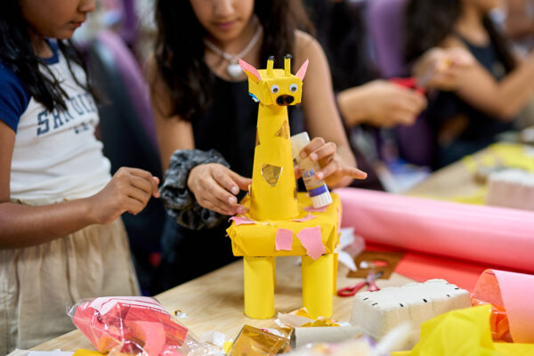 A giraffe model crafted from yellow paper and google eyes is glued together by two young children