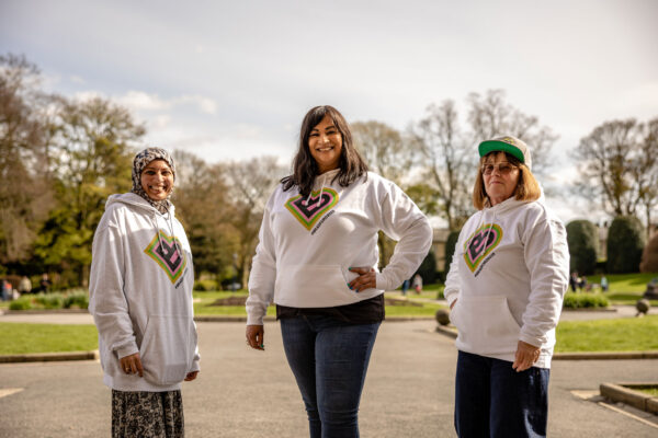 Three women pose in a park wearing white hoodies with the Bradford 2025 logo on the front