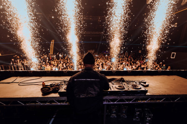 Image shows a DJ from behind, performing in front of a crowd. Pyrotechnics explode in front of the stage.