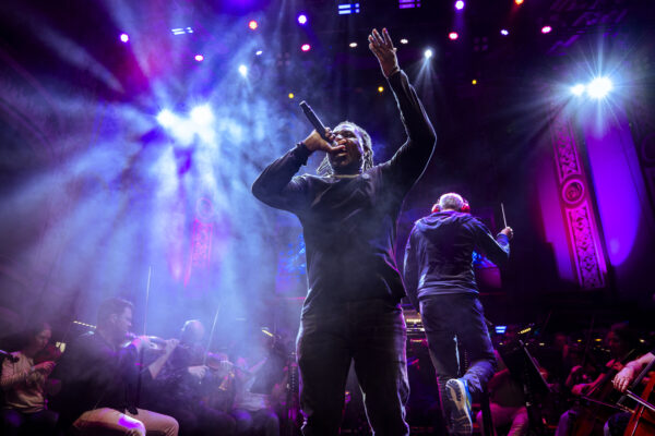 A black man speaks into a microphone on stage - the orchestra is behind him.