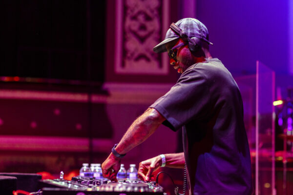 A black man works on a DJ deck - he is wearing a hat and headphones, as well as a dark t-shirt.
