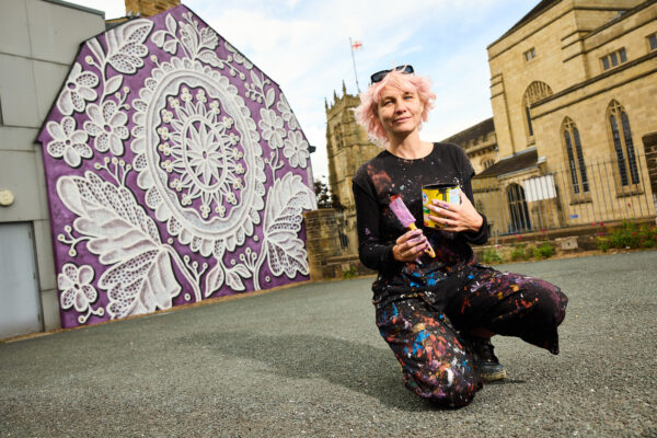 Artist, Nespoon knelt in the foreground with paint brushes in her hand, she has paint splashes on her knees. Behind her is the mural, which is a large white lace design that sits on a purple background.