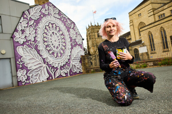 Artist, Nespoon, is crouched in front of the mural she has painted, with a paintbrush in hand. The mural is a white lace pattern that sits on a purple background. There is a historic building in the corner of the image.