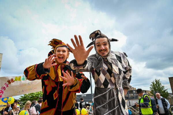 Two performers in vibrant costumes wave towards the camera.