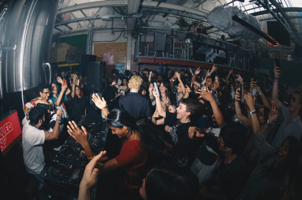 A crowd of people with their hands in the air in a dimly lit room, dancing to a DJ set.