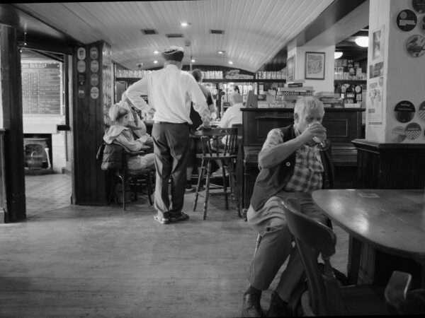 Black and white image of a group of people in a pub. A man sitting close to us is drinking a pint of beer.