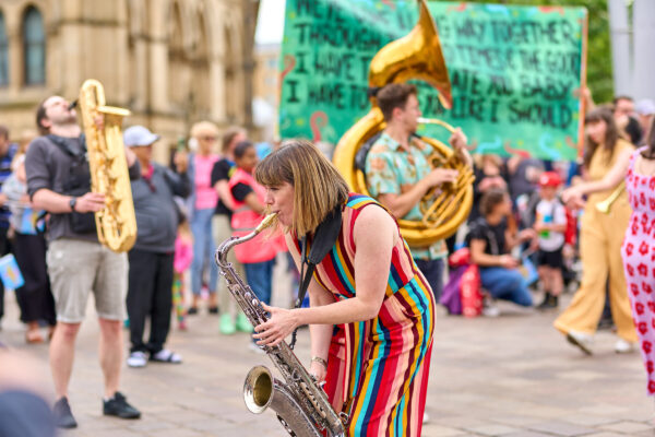 A musician plays a saxophone in the street. They are surrounded by other musicians playing brass instruments.