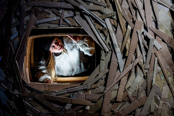 A young woman in a white hoodie and black beanie hat looks through a window in structure made out of cardboard slats.