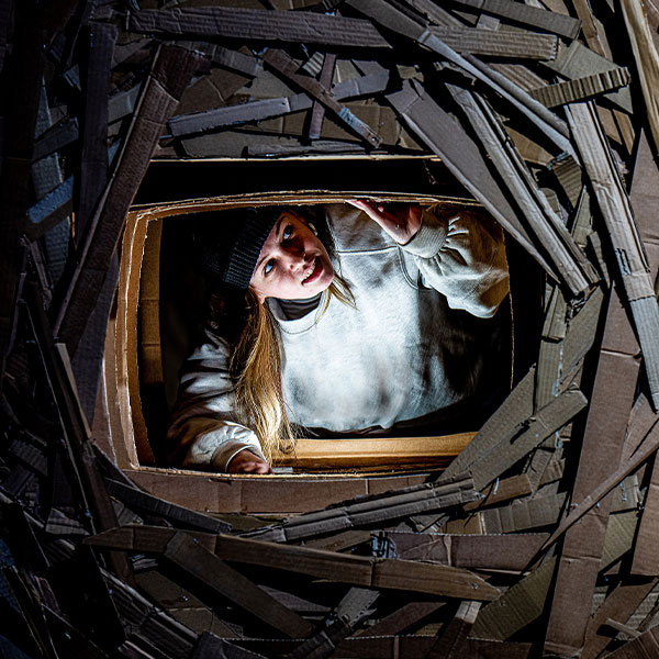 A young woman in a white hoodie and black beanie hat looks through a window in structure made out of cardboard slats.