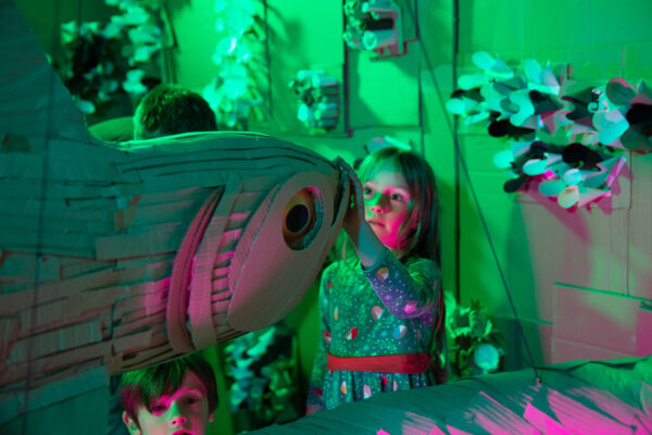 A young girl in a flowery dress puts her hand on the mouth of a cardboard fish. A young boy is in the bottom left of the image.