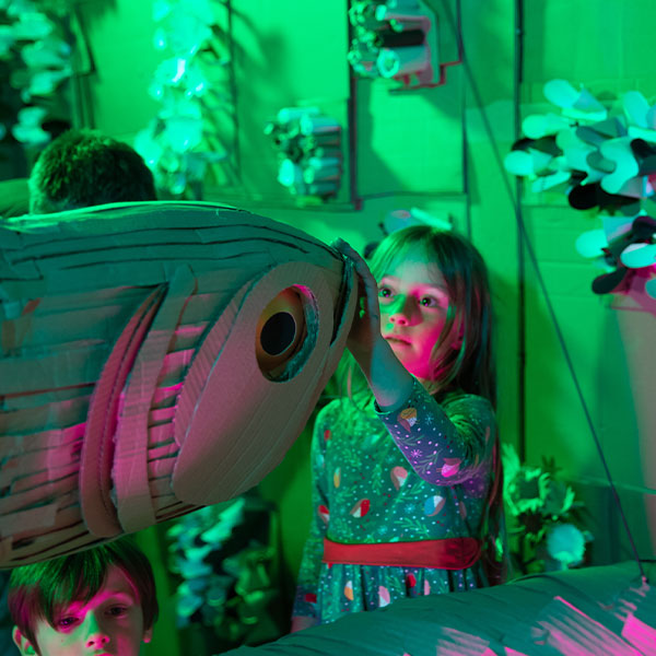 A young girl in a flowery dress puts her hand on the mouth of a cardboard fish. A young boy is in the bottom left of the image.