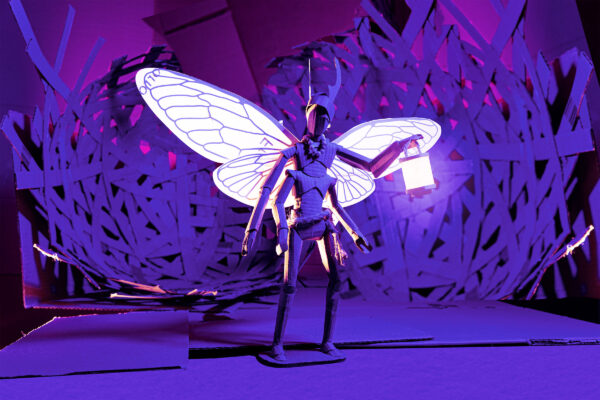 A bug holding a lantern with lit up wings against a forest style backdrop, all of which is made out of cardboard