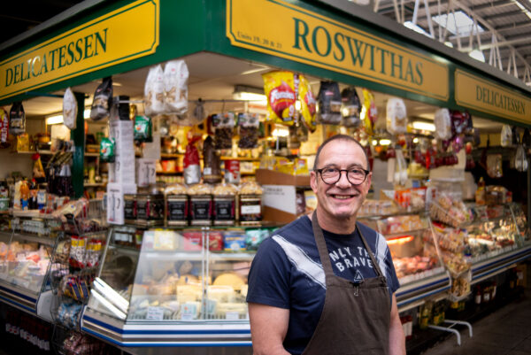 A man wearing an apron stands in front of a deli counter.