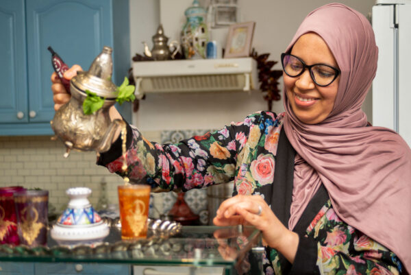 Mariem pours tea from a metal pot. She wears a hijab and is smiling.