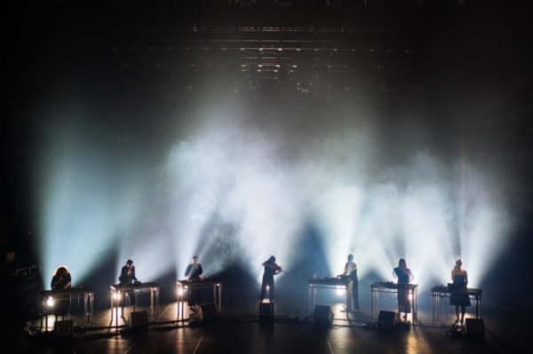 A group of musicians performing on stage. They are all stood in a line silhouetted by white light behind them.