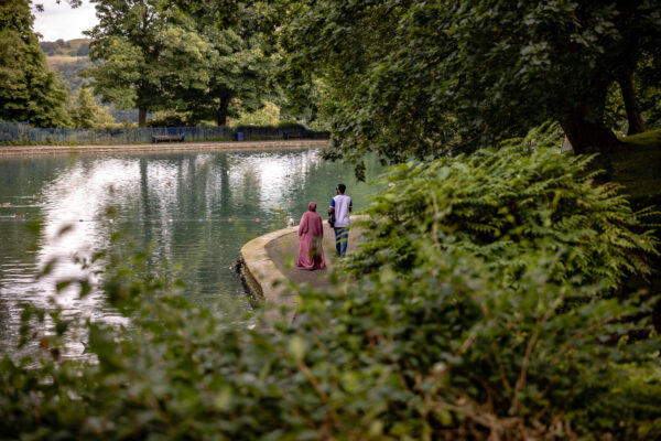 A lake can be seen in the left hand side of the image, with green foliage in the foreground of the image. Two people are walking together round the lake on the path.