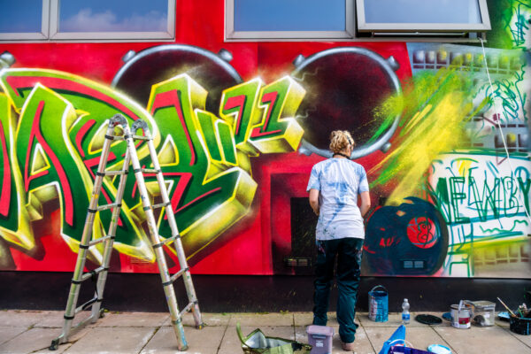 Yellow and green spray painted words 'MAPA', sitting on a red background. There is a ladder to the left of the picture and artist, Peachzz with her back turned to the right.