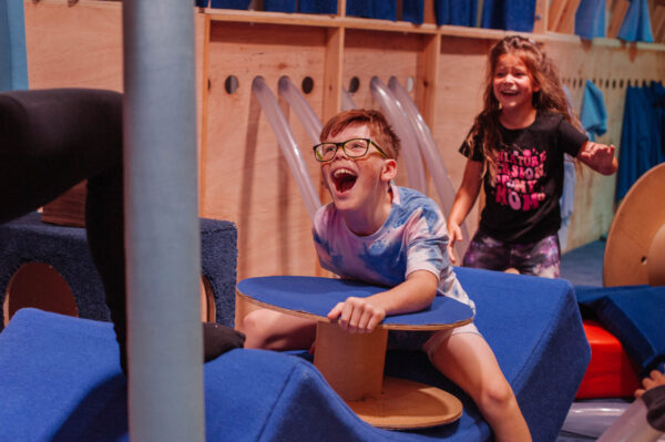 Two young people playing on a large bobbin structure. They are laughing.