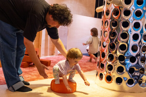 A man leans down to guide a toddler in a soft play area